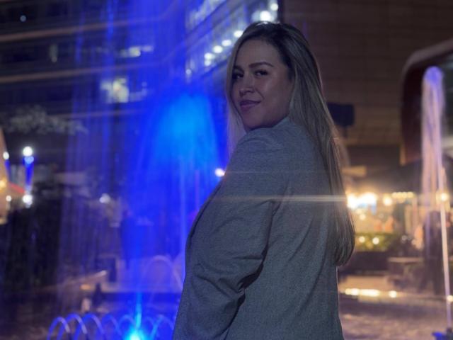 LiaDiane, com seios médios, sorrindo, olhando por cima do ombro, camisa cinza de manga comprida, cabelo loiro, ambiente noturno, luz azul, reflexos de água, leve efeito de lente, confiante, atraente..