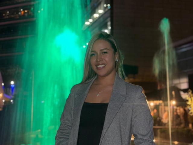 LiaDiane, con i capelli biondi, sorridente, indossa un blazer grigio e una maglietta nera, sullo sfondo c'è una fontana d'acqua verde, appare felice e sicura di sé, di corporatura media, carina..