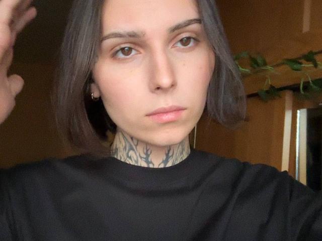 A TransWithLove tem pele clara, cabelo castanho na altura dos ombros, tatuagens no pescoço, olhos castanhos e veste uma camisa preta, com uma expressão séria. Suas mãos estão parcialmente levantadas..