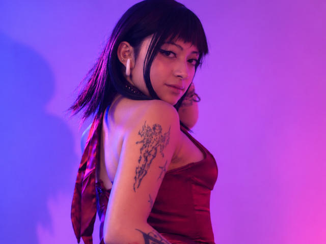 DarlingKeit with purple-black hair, red halter top, tattoos on shoulder and arm, looking back, pink-blue lighting, small earrings, soft smile, sexy, cute, attractive.