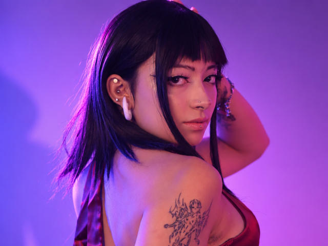 DarlingKeit with black hair, purple-blue lighting, red lace bra, nose ring, white earbuds, tattoo on right shoulder, looking back, hand in hair, sexy, cute, attractive.