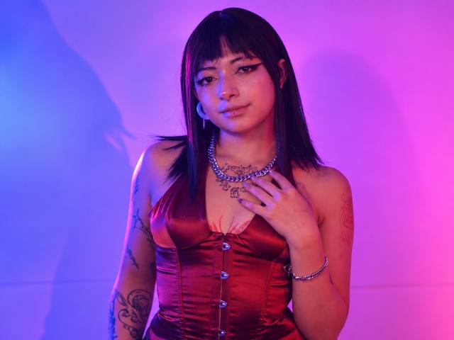 DarlingKeit with black hair, bangs, and tattoos, wearing a red satin corset with buttons, silver necklace, and bracelet, under purple and blue lighting. Boobs are visible.