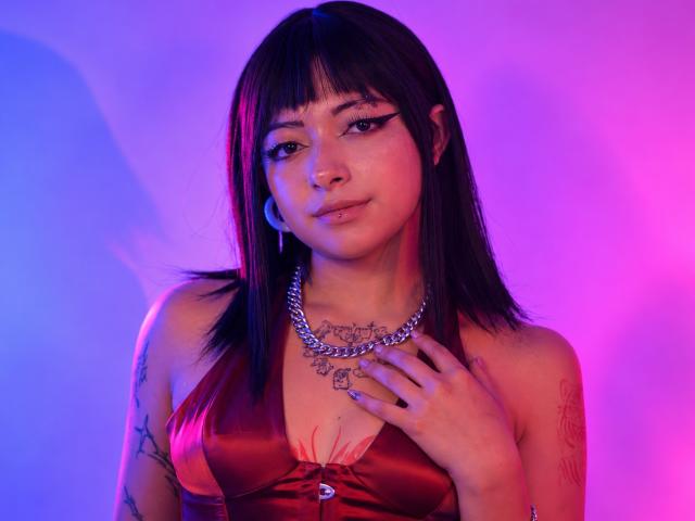 DarlingKeit with straight black hair, nose ring, and eyeliner, wearing a red satin top with a chain necklace, holding her chest, tattoos on arm, pink and blue lighting.