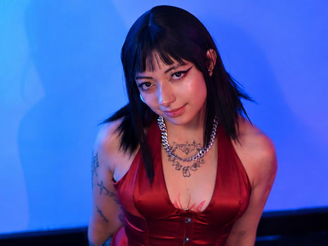 DarlingKeit with black hair, red satin top, and silver chain, showing off tattoos and a cute smile, her tits barely covered, in a sexy, confident pose.