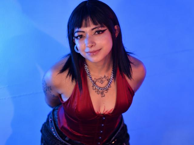 DarlingKeit with black hair, red velvet top, and necklace, looking up with a smile, showing her tits, pierced nose, and tattoos, wearing dark pants, in blue light.
