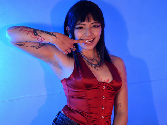DarlingKeit with dark hair, red satin corset, tattoos, chain necklace, and blue lighting, pointing at her smiling mouth with a finger, showing her tits and armpit hair.