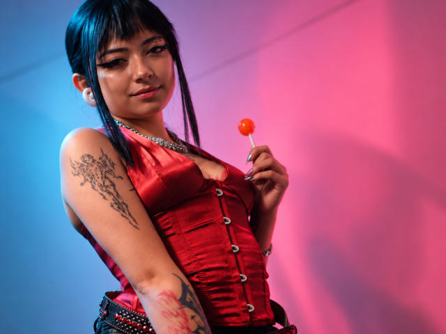 DarlingKeit with black hair, bangs, and tattoos, holding a red candy, wearing a red satin corset, silver necklace, and black belt, against a pink and blue light.