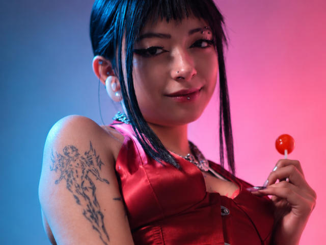 DarlingKeit with black hair, nose ring, and eyeliner, holding a red candy, wearing a red satin top, showing a tree tattoo on her left arm, smiling.
