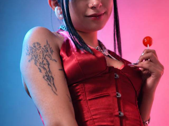 DarlingKeit with dark pigtails, holding a red lollipop, wearing a shiny red button-up top, showing a tattoo on her right shoulder, and a nose ring. Boobs visible.