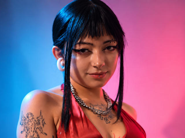 DarlingKeit with black bangs, nose ring, and eyeliner, wearing a red satin halter, silver chain, and white earrings, showcasing tattoos on her shoulder and chest, smiling subtly.