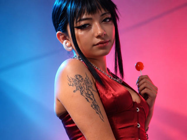 DarlingKeit with black hair, wearing a red satin dress, holding a red lollipop, has a tattoo on her right arm, and a nose ring, against a pink-blue gradient.