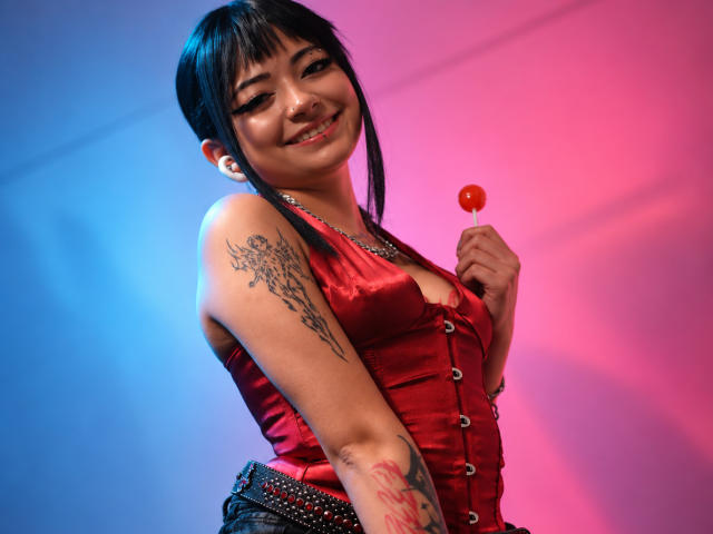 DarlingKeit with black hair, red satin top, holding red candy, smiling, tattoo on right arm, earrings, and belt with studs, pink-blue lighting, looking up at camera.