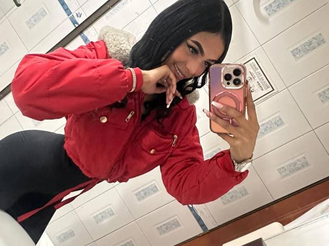 TatianaLangley with long black hair, smiling, wearing a red jacket and black skirt, taking a mirror selfie, showing off her curvy ass, holding a pink phone.