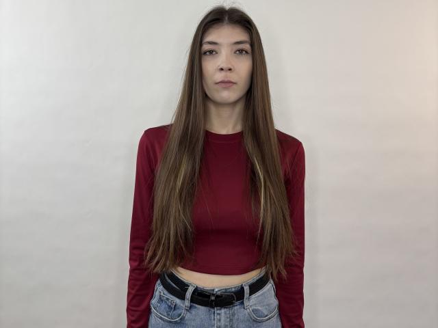 InaSunderlin tem cabelos castanhos longos, veste uma blusa cropped vermelha de manga comprida e jeans azuis com um cinto preto, e está em pé em frente a uma parede branca lisa..