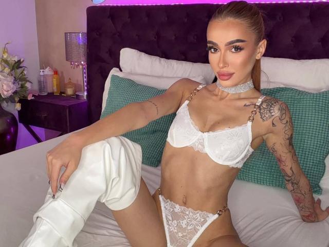 MarissaFex mit Tattoos, trägt einen weißen Spitzen-BH und String, weiße kniehohe Stiefel, eine Halskette und einen Pferdeschwanz, sitzt auf dem Bett, zeigt kleine Brüste und einen schlanken Hintern, mit violettem Licht..