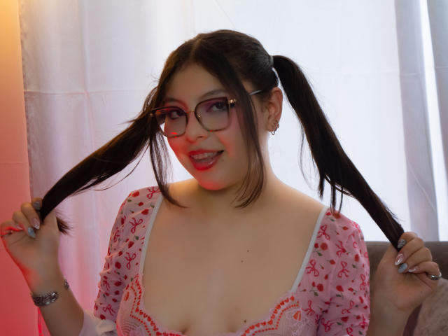 AnnaBeltrann, avec des couettes, des lunettes, un haut rose à motifs floraux, dévoilant sa poitrine, souriante, se tenant les cheveux, rouge à lèvres, bracelet argenté et boucles d'oreilles. Ses seins sont petits et elle est mignonne..