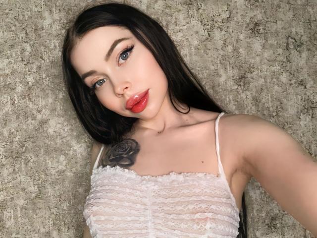 VelvetD with long black hair, red lips, and white lace top showing her small tits and rose tattoo on her right shoulder. She has blue eyes and a cute nose.