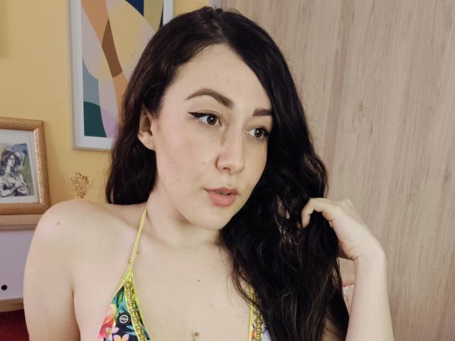 Maby Legrand, com longos cabelos pretos, vestindo um top colorido, tem uma expressão de lábios grossos, segura o cabelo e tem seios pequenos. Pele clara, delineador de olhos e uma pequena perfuração no nariz..