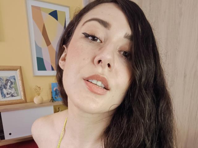 El rostro de Maby Legrand, piel pálida, cabello oscuro y ondulado, delineador de ojos negro alado, labial rosa suave, labios rosados ligeramente separados, collar dorado, pared amarilla, puerta de madera en el fondo..