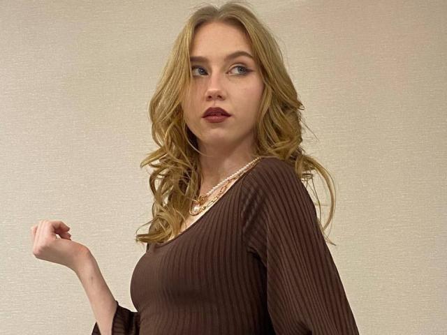 ShawtyOana has blonde wavy hair, blue eyes, and is wearing a brown ribbed top with a pearl necklace, looking slightly to the side with a neutral expression.