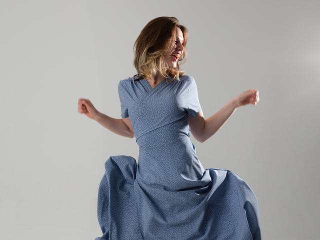 MicheleIreland is jumping with arms raised, wearing a blue, short-sleeved, polka-dotted dress that flares out, showing a playful, joyful expression, and blonde, wavy hair.