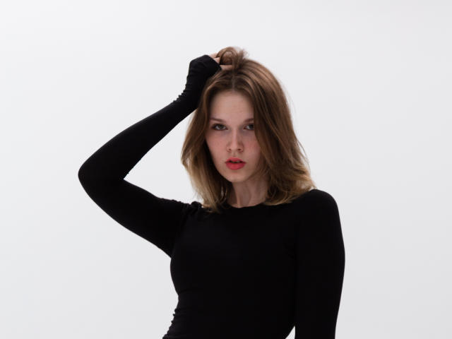 MicheleIreland with medium tits, wearing a black long-sleeve top, has her hand in her brown hair, looking serious with red lips, against a white background.