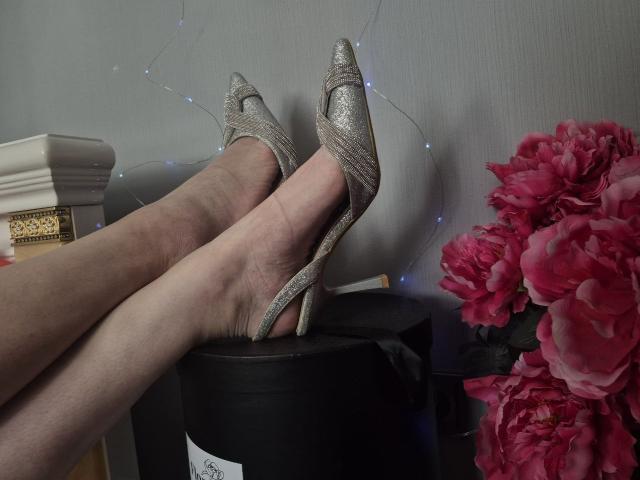 WenTina's legs in glittery silver high heels, crossed at the ankles, resting on a black stool. Pink peony flowers on the right, blue fairy lights in the background.