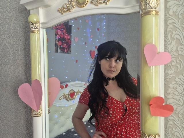 WenTina with black hair, wearing a red polka dot dress, choker, looking in a mirror with pink and red heart decorations, hands on hips, medium tits, bedroom setting.