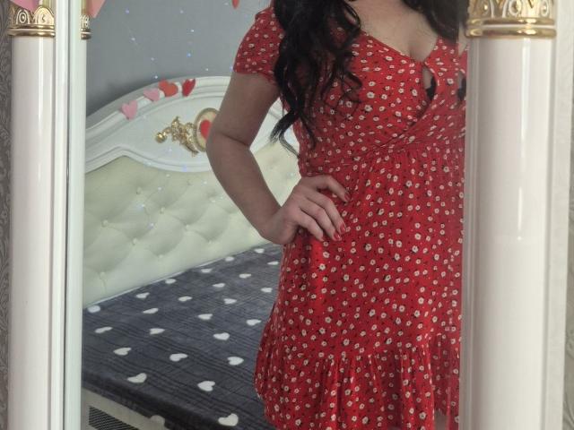 WenTina, con cabello negro largo, vestida con un vestido rojo con pequeños patrones blancos, de pie frente a un espejo, con una mano en la cadera, mostrando sus pechos y un ligero escote..