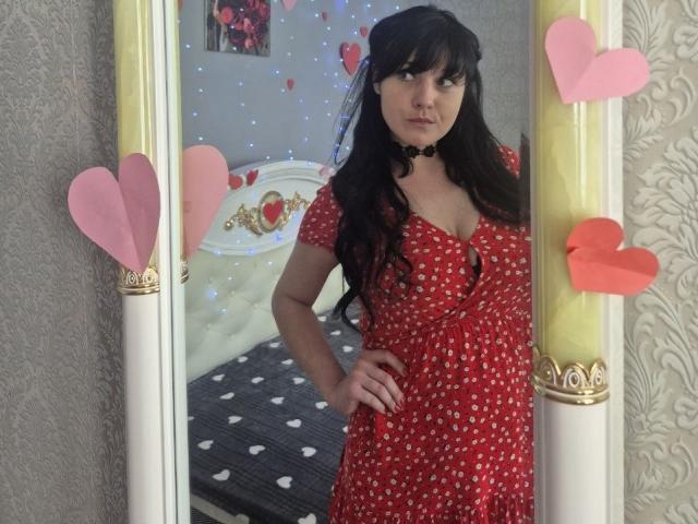 WenTina, com cabelos pretos longos, um vestido vermelho com bolinhas pretas e um colar preto, está em frente a um espelho com recortes em formato de coração rosa e vermelho. Ela está olhando para a câmera..