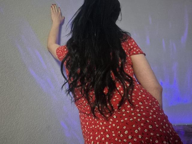 WenTina, com longos cabelos pretos ondulados, vestindo um vestido vermelho com padrões brancos, em pé contra uma parede, com a mão na parede, nádegas levemente elevadas, luz azul à direita..