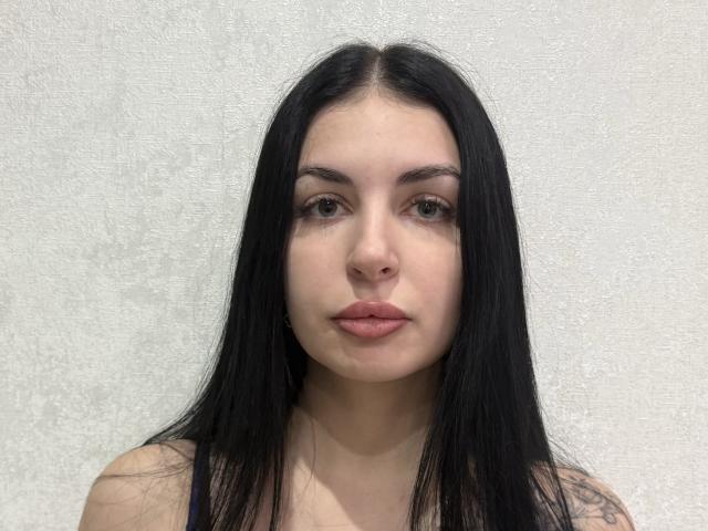 JustineGreaser tiene el pelo largo y negro, piel clara, ojos marrones y labios rosados. Lleva una camiseta oscura sin mangas y mira directamente a la cámara con una expresión neutra..