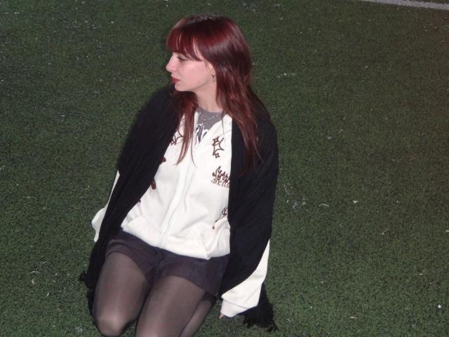 LeomaHaq with red hair, black cardigan, white shirt, brown shorts, and black tights, kneeling on grass, looking to the side, showing her ass slightly.