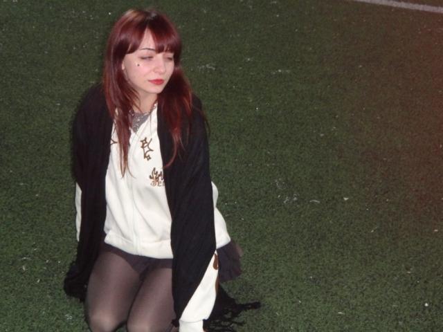 LeomaHaq with red hair, black shawl, white top, black pantyhose, and black underwear, kneeling on green grass, eyes closed, red lipstick, small black mole on left cheek.
