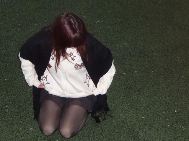 LeomaHaq, kneeling on grass, wearing a white and black jacket, black shorts, and black pantyhose, with her back to the camera, brown hair covering her face.
