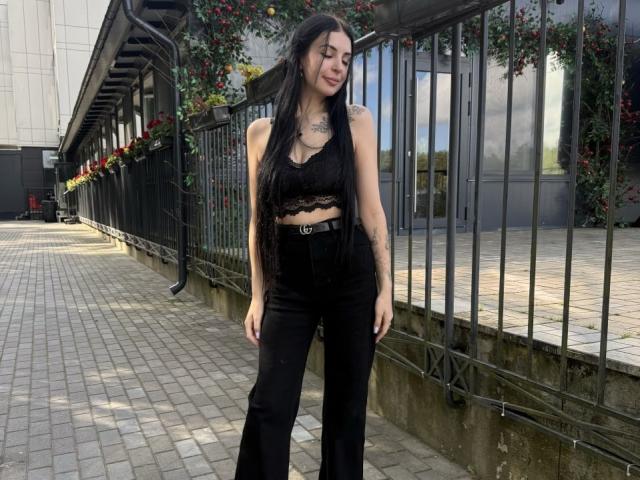 JustineGreaser mit langem, schwarzem Haar, kleinen Brüsten, einem schwarzen Spitzen-Crop-Top, einer schwarzen Hose mit hoher Taille, lächelnd, stehend auf einem Ziegelweg, ein schwarzer Metallzaun mit roten Blumen im Hintergrund..