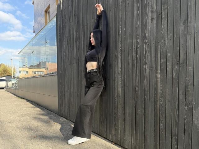 JustineGreaser con sudadera negra con capucha, top corto negro, pantalones negros de cintura alta y zapatillas blancas, con los brazos levantados, apoyada contra una pared de madera oscura, mostrando sus abdominales planos y sus zapatillas blancas..