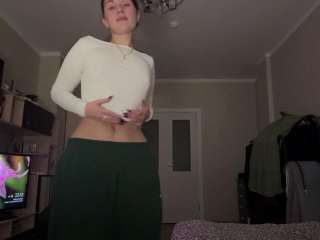Marva Oberdorf está de pie en una habitación, vestida con una camiseta blanca de manga larga y pantalones verdes, con las manos sobre su abdomen, mostrando su vientre delgado y plano. Lleva un collar y tiene el pelo corto y castaño..