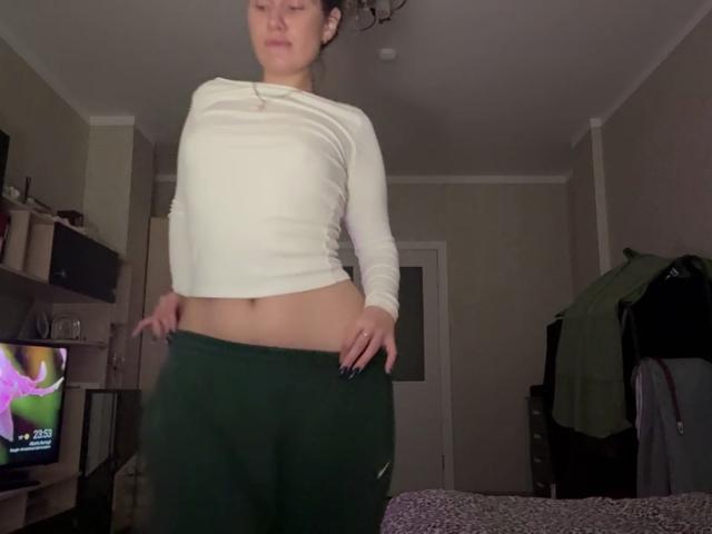 Marva Oberdorf staat in een kamer, gekleed in een witte blouse met lange mouwen en groene broek, met haar handen op haar heupen, waarbij een klein deel van haar platte achterwerk en haar blote buik zichtbaar zijn..