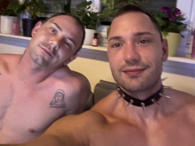 Deux Twunkis sans chemise, avec la tête rasée, dont l'un a un tatouage de fantôme sur la poitrine et un collier clouté, et l'autre qui sourit légèrement. Tous deux ont une peau claire et une musculature développée..