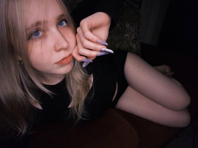 TiffaniMacandog, avec ses cheveux blonds, ses yeux bleus et ses ongles violets, est allongée sur un canapé rouge, en sous-vêtements noirs, montrant ses cuisses et mordant sa doigt..