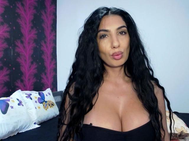 BrunetteBabe69 con cabello negro largo y ondulado, labios rosados y voluptuosos, y grandes senos, vestida con un top negro sin tirantes, sentada en una cama con almohadas de colores..