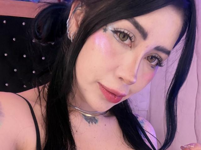 Kkole with black hair, green eyes, and pink blush, wearing a choker and black straps, has a tattoo on her neck and a shiny earring, looking at the camera.