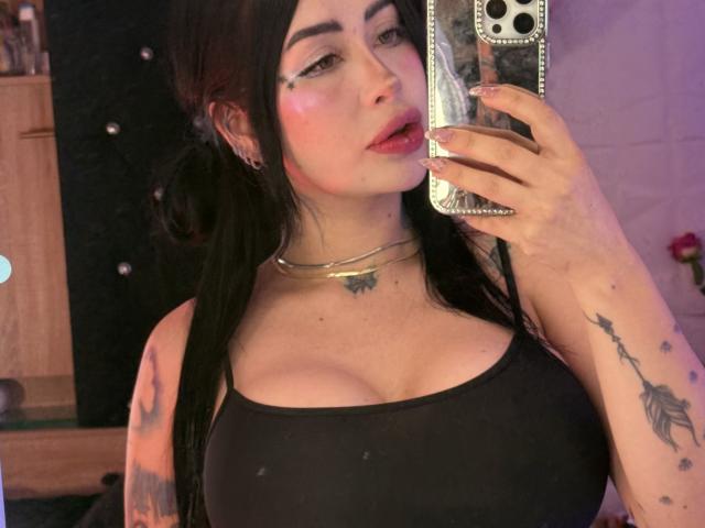 Kkole with black pigtails, wearing a black tank top, showing big tits, tattoos on arms, holding phone, gold necklace, pink blush, looking at mirror selfie.