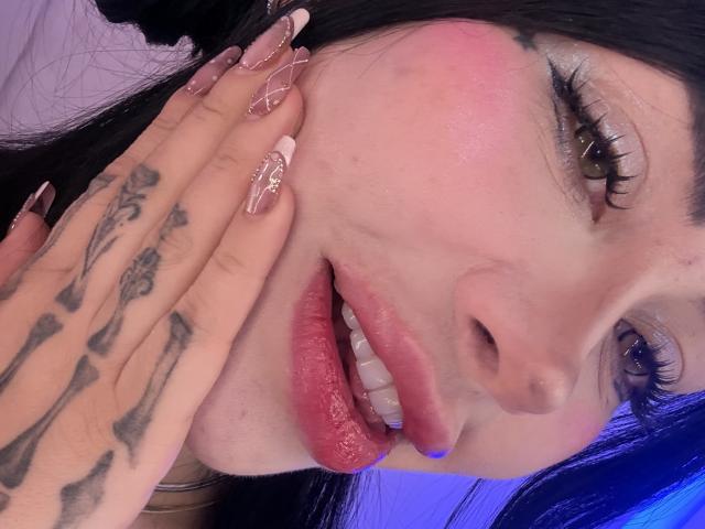Kkole's face, pink blushed cheeks, brown eyes, black hair, glossy pink lips, hand with bone tattoos, long pink nails, slight smile, blue light background.