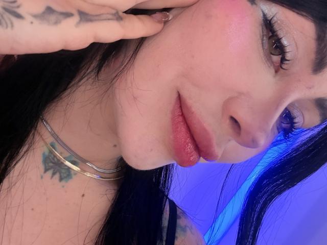 Kkole's face with pink blush, full pink lips, and brown eyes. She has black hair, a tattoo on her neck, and a tattoo on her hand. Blue light background.