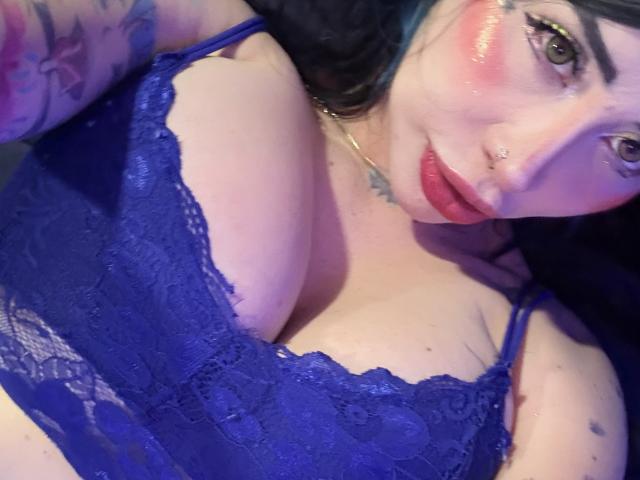 Kkole with blue lace bralette, showcasing her tits, pink blush on cheeks, nose ring, tattoos on arm, and red lipstick, taking a selfie with a slight downward angle.