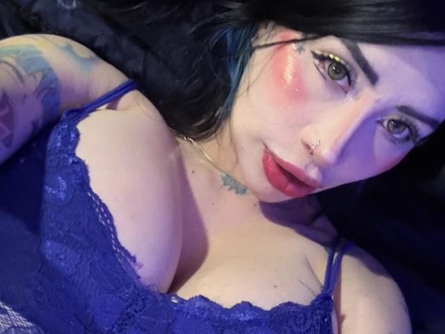 Kkole with pink blushed cheeks, black hair, nose ring, and blue lace bra, showing large tits, tattooed arms, and green eyes. Her lips are slightly parted.