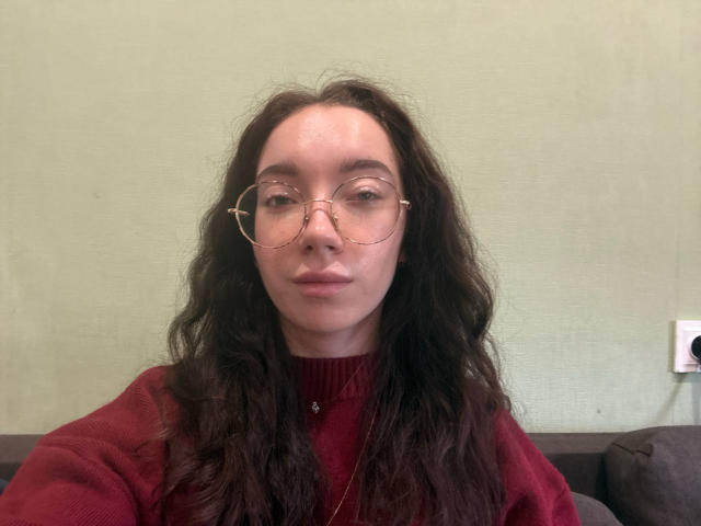 Claire Spisak, mit welligem, dunkelbraunem Haar, trägt eine runde, goldfarbene Brille und einen roten Pullover. Sie hat einen neutralen Gesichtsausdruck und einen kleinen Nasenring in ihrem linken Nasenloch..