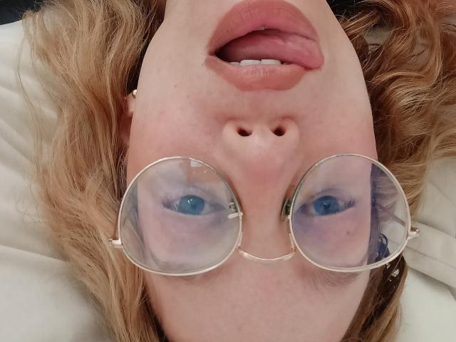 MissFrenchy's face, looking up with her tongue slightly out, wearing round glasses, showing blue eyes, and fair skin with light brown hair. Her pink lips are slightly parted.